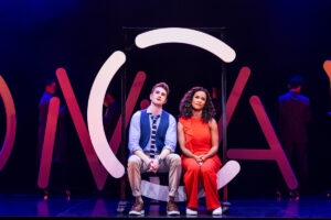 David Socolar as Theo and Britney Coleman as Bobbie sitting on a neon C, in the North American Tour of COMPANY. Photo by Matthew Murphy for MurphyMade